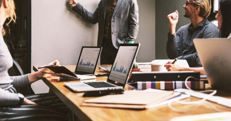 "Team meeting in a modern office setting with laptops displaying financial charts, documents, and a presenter explaining data insights on a whiteboard, symbolizing effective project cost management."